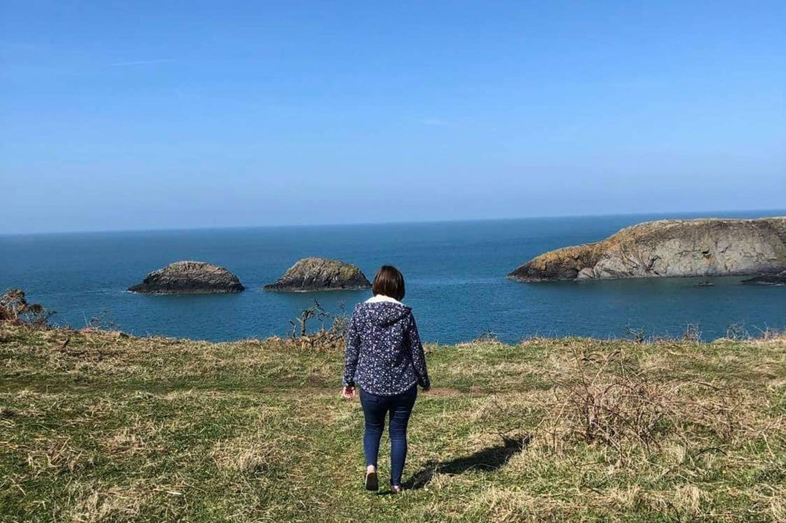 A photo of a girl (Chloe from Chloe Evans Designs) walking along the Welsh Coastal Path with islands, blue sky, and sea in the background.