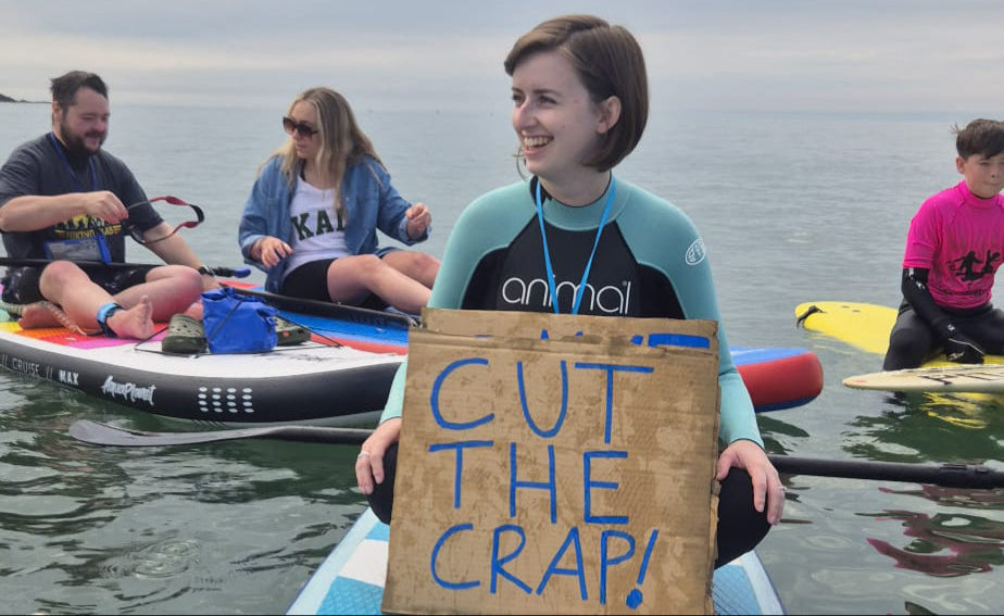 A photo of Chloe from Chloe Evans Designs on a paddleboard holding a 'Cut the crap' placard at a Surfers Against Sewage protest in 2024.