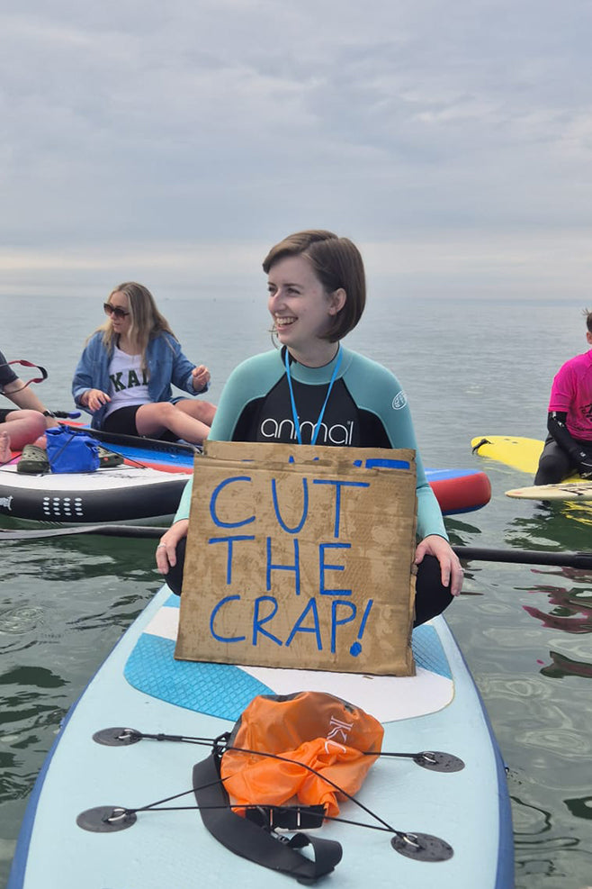 A photo of Chloe sat on her paddleboard with a 'Cut the Crap' placard at a Surfers Against Sewage protest at Caswell Bay in Gower.