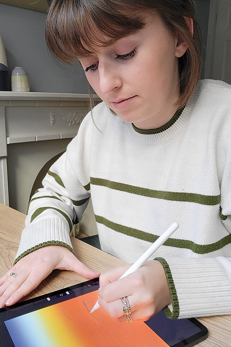 A photo of Chloe sat at her desk, drawing a new sunset design on her iPad.