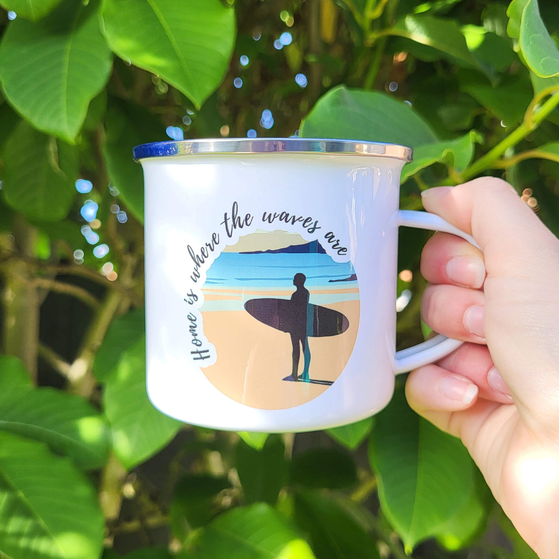 White enamel mug showing surfer looking out to sea, photographed outside in natural light.