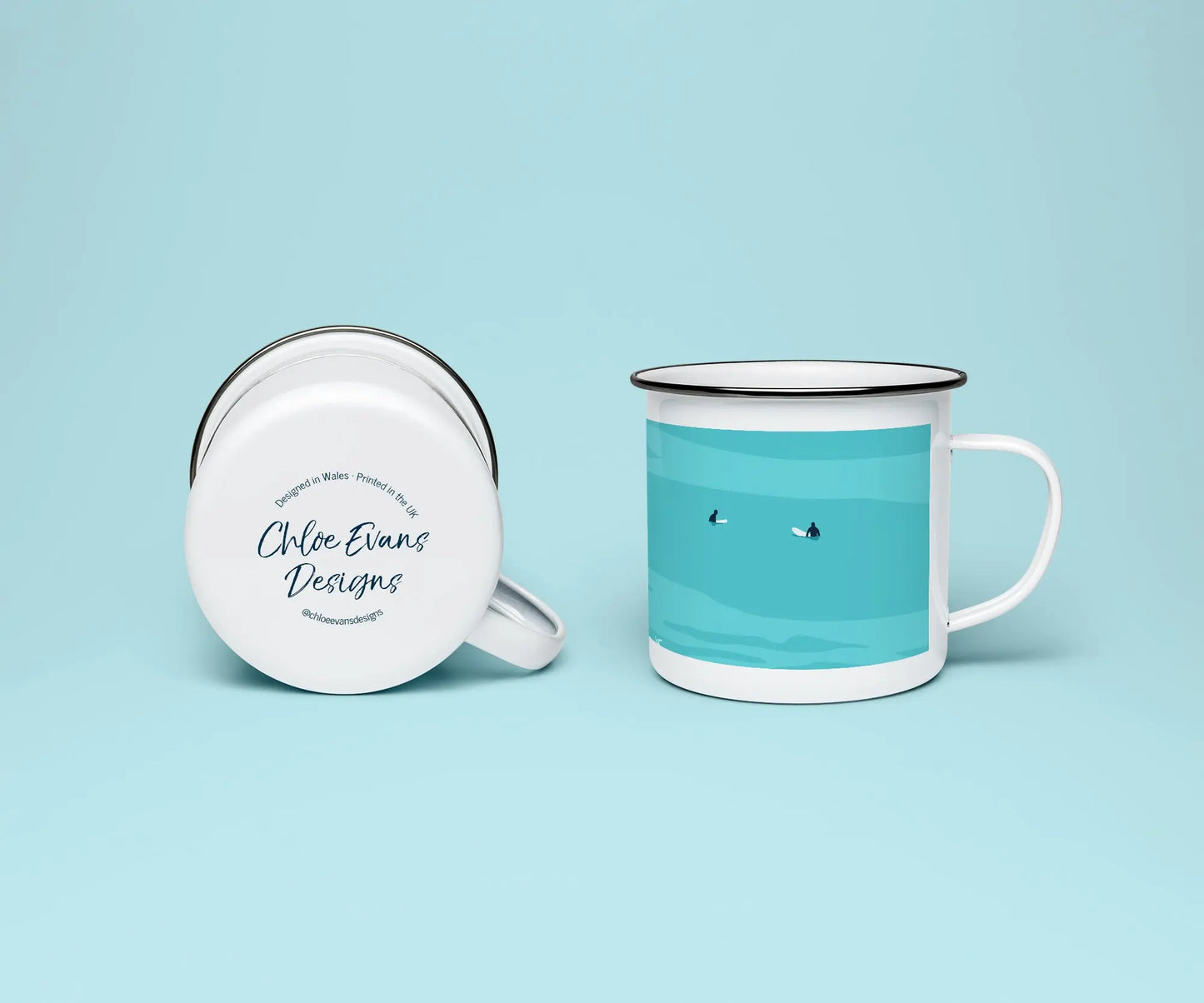 A white enamel mug from two perspectives: one of the mug on its side showing the branded base stamp on the bottom of the mug, and the other of the mug upright showing the blue print of two surfers; all on a light blue background.