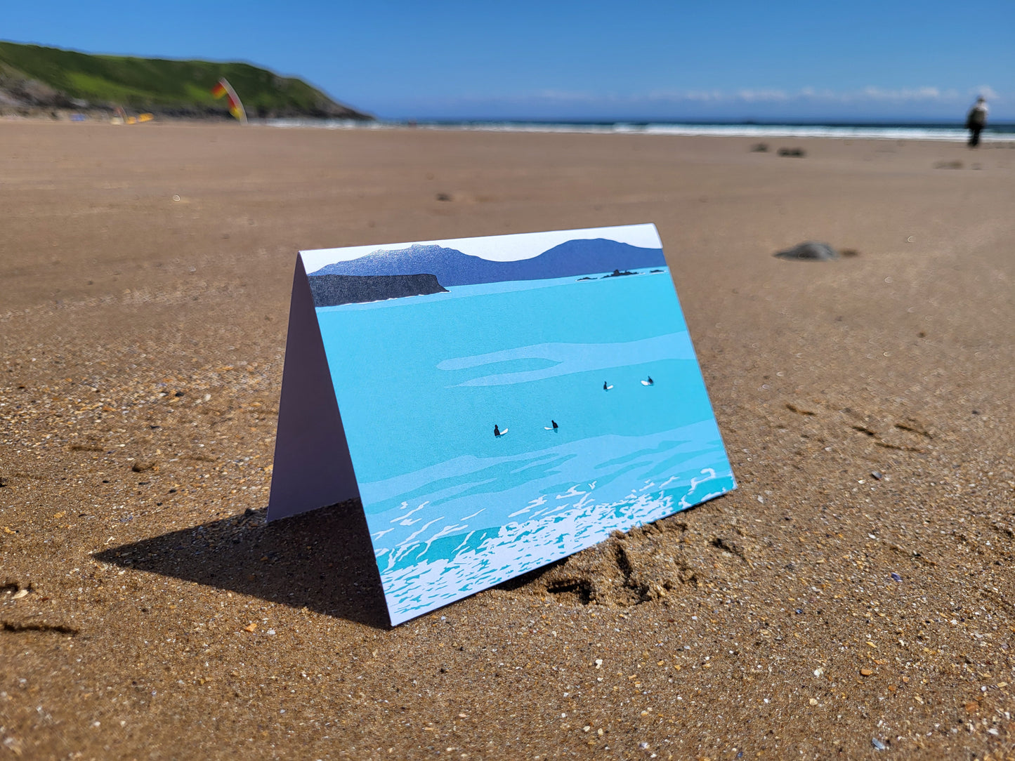 A photo of a greeting card with an surf inspired design, pictured on the beach.