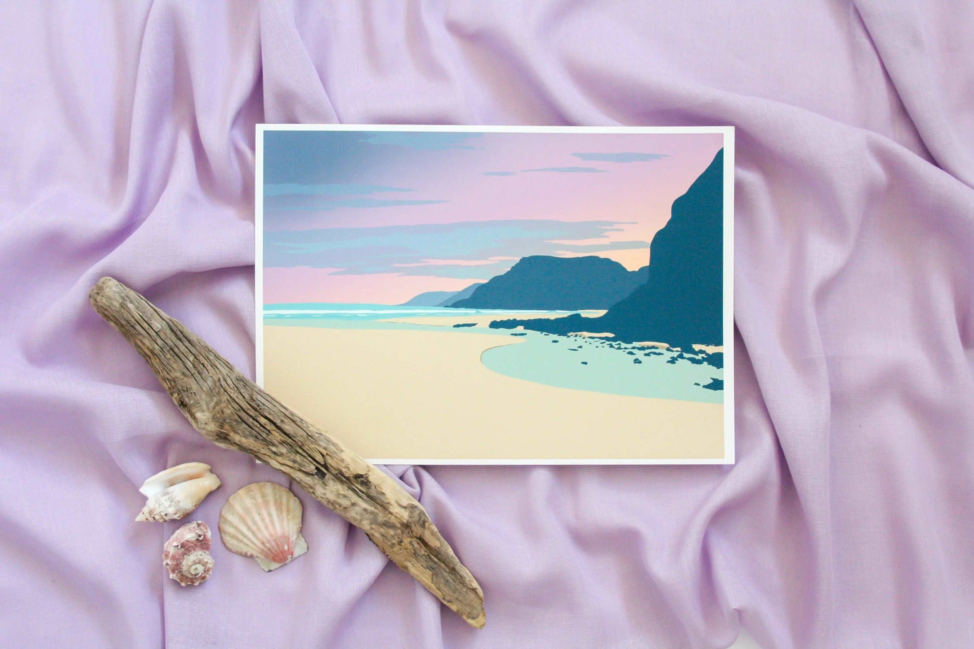 Pink and blue Caswell beach sunset print on pink fabric backdrop with driftwood and shells props.