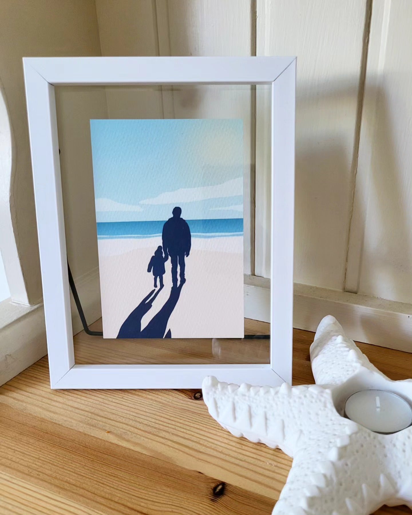 A photo of a custom illustration of a father and sun holding hands on the beach, in a white photo frame, on a welsh dresser with a starfish shaped tealight holder next to it.