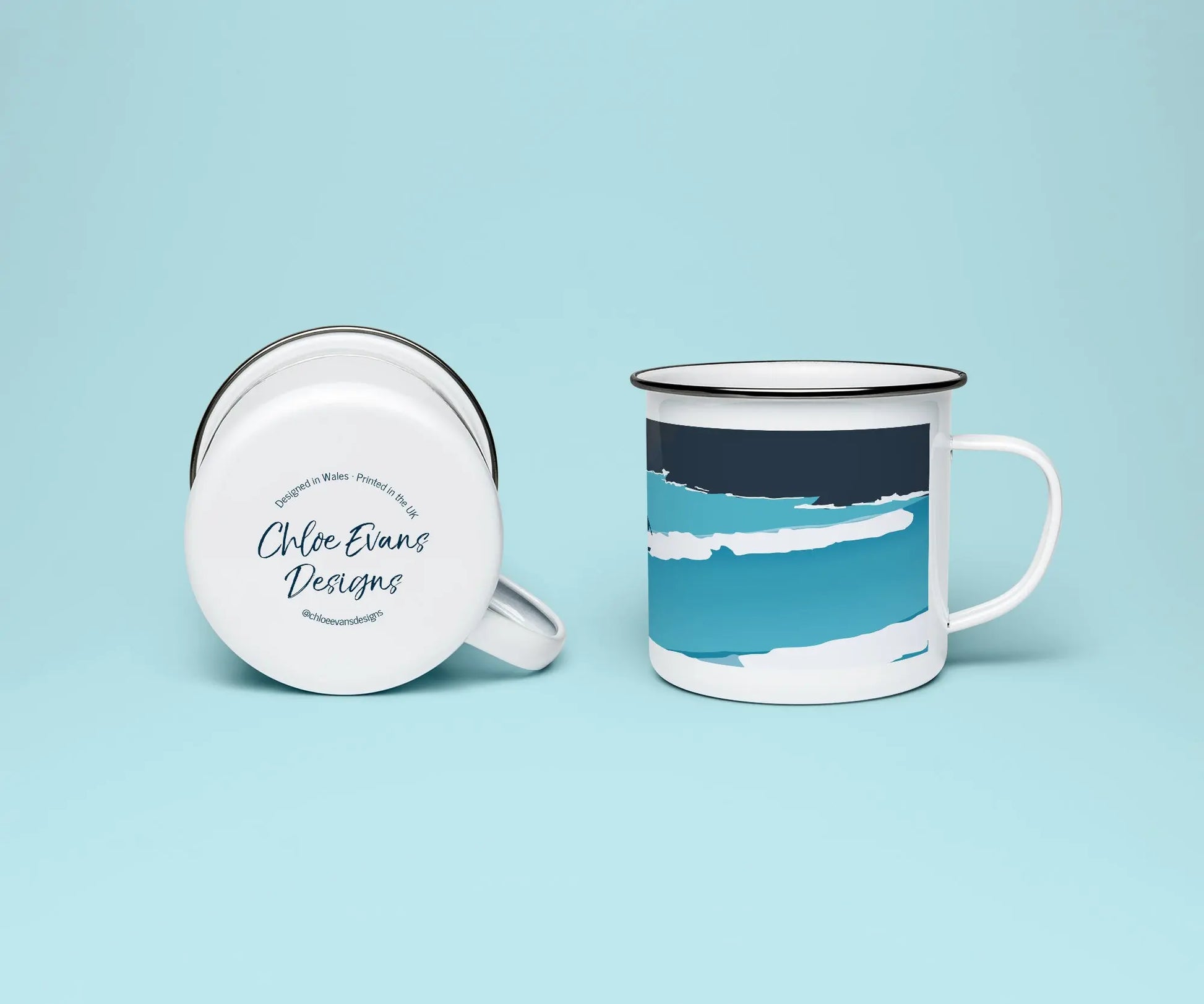 A white enamel mug from two viewpoints, one upright showing it's blue print of a wave, and the other on it on its side showing the branded base stamp on the bottom of the mug; all on a light blue background.