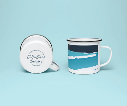 A white enamel mug from two viewpoints, one upright showing it's blue print of a wave, and the other on it on its side showing the branded base stamp on the bottom of the mug; all on a light blue background.