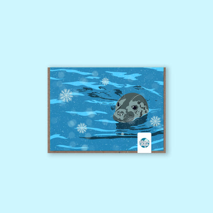 A greeting card featuring an illustrated grey seal sitting in falling snow, with a wintry coastal background.