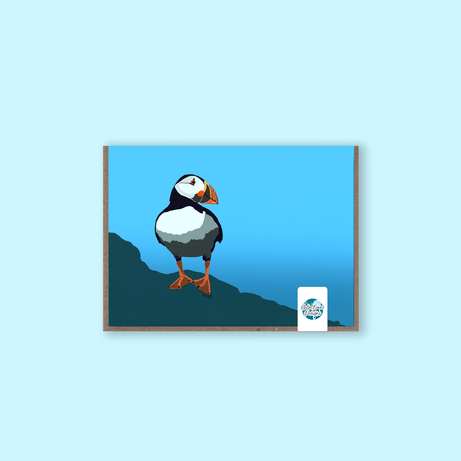 A mockup of a greeting card with an illustration of a puffin on the front, with a brown envelope behind and plastic-free removable sticker packaging, on a light blue background.