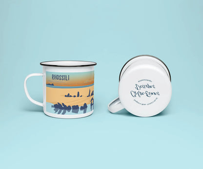 A white enamel mug from two perspectives: one of the mug on its side showing the branded base stamp on the bottom of the mug, and the other of the mug upright showing the blue and orange illustration of the Helvetia shipwreck on Rhossili beach at sunset; all on a light blue background.