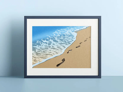 A mock up of a print of footprints in the sand along the shore line, disappearing into the distance. The print is frames and displayed against a light blue background.