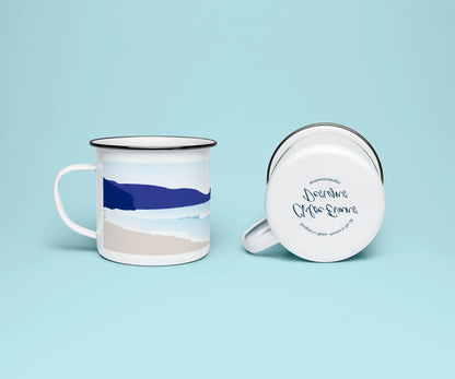 A white enamel mug from two perspectives: one of the mug on its side showing the branded base stamp on the bottom of the mug, and the other of the mug upright showing the blue print of Newgale beach; all on a light blue background.