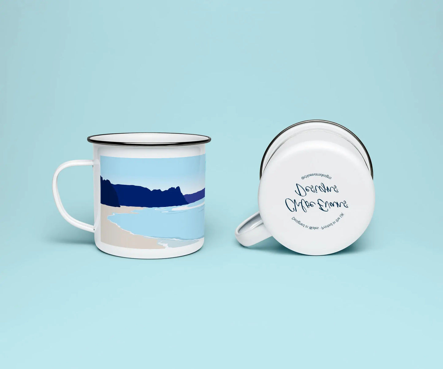 A white enamel mug from two perspectives: one of the mug on its side showing the branded base stamp on the bottom of the mug, and the other of the mug upright showing the blue print of Oxwich beach; all on a light blue background.