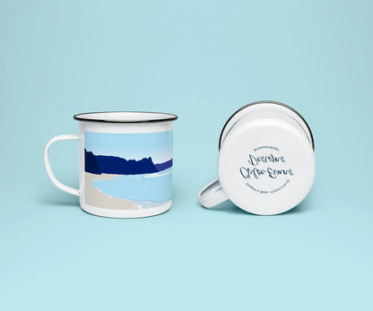 A white enamel mug from two perspectives: one of the mug on its side showing the branded base stamp on the bottom of the mug, and the other of the mug upright showing the blue print of Oxwich beach; all on a light blue background.