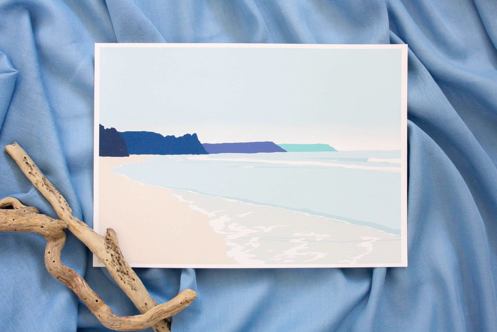 Photo of blue and turquoise Oxwich bay illustration print on blue fabric backdrop and driftwood