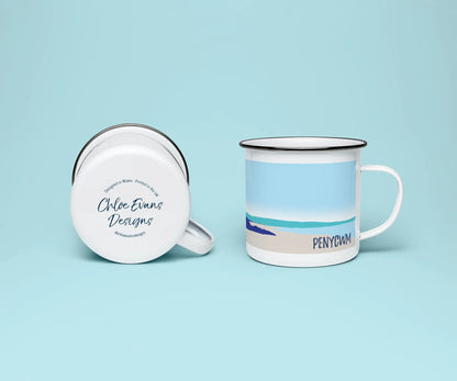 A mock up of a white enamel mug from two perspectives; one of the mug on its side looking at the bottom of the mug and the branded base stamp, and the other of the mug upright showing the print of an orange and blue beach scene, showing the rugged coastline at Penycwm beach, all on a light blue background.