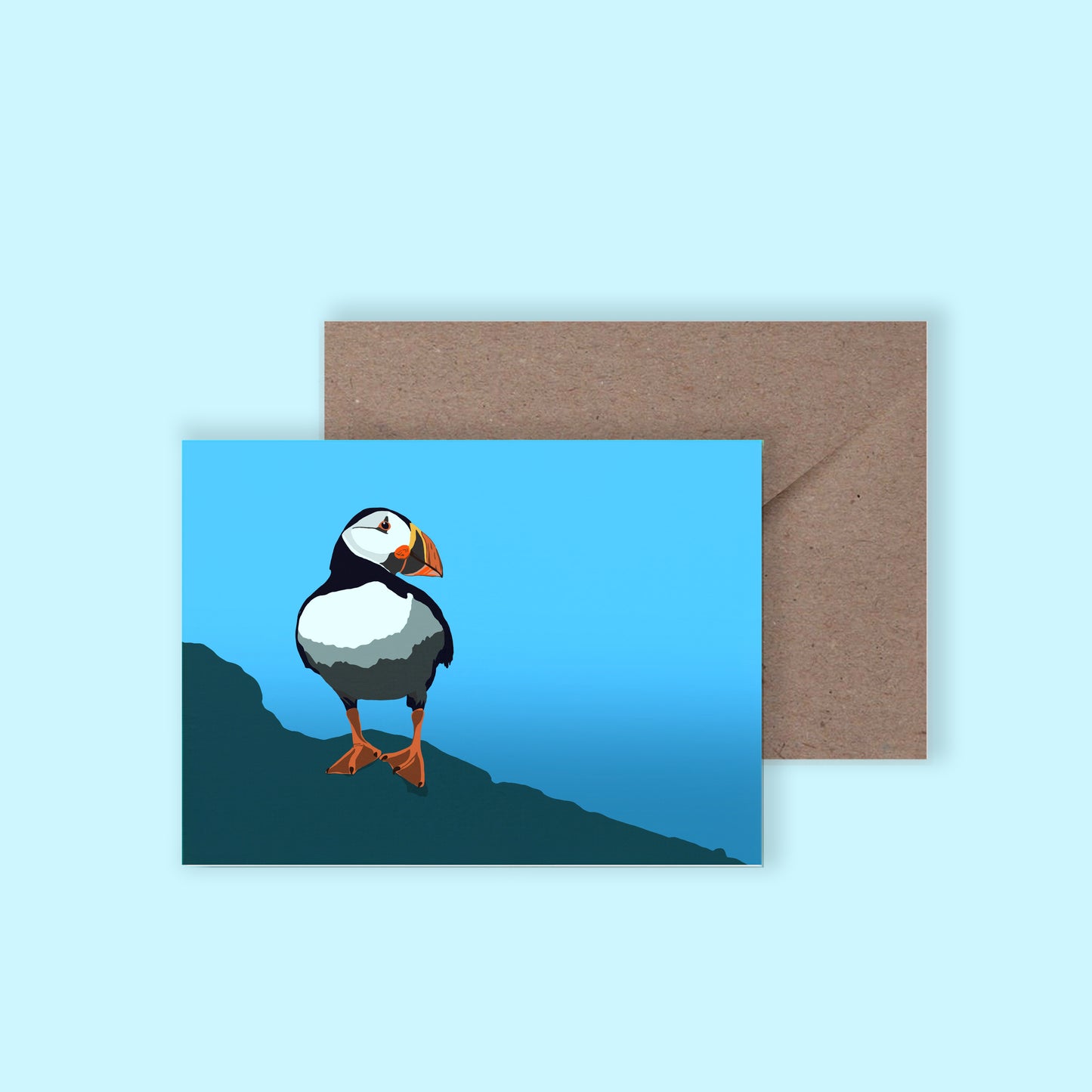 A mockup of a greeting card with an illustration of a puffin on the front, with a brown envelope behind, on a light blue background.