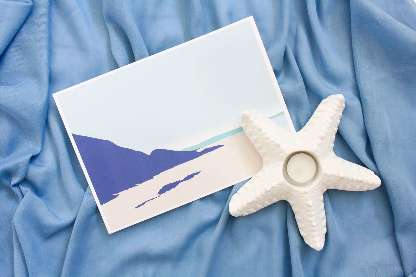 Photo of Pwll March to Newgalde beach illustration print on blue fabric backdrop and starfish tealight holder decoration