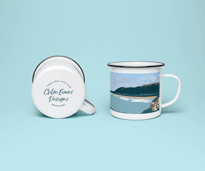 A white enamel mug from two perspectives: one of the mug on its side showing the branded base stamp on the bottom of the mug, and the other of the mug upright showing the green and blue print of Rhossili beach; all on a light blue background.