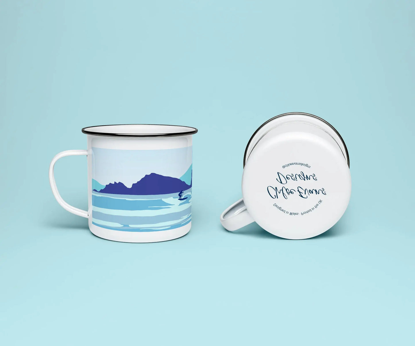 A white enamel mug from two perspectives: one of the mug on its side showing the branded base stamp on the bottom of the mug, and the other of the mug upright showing the blue print of a paddleboarder surfing with cliffs in the background; all on a light blue background.