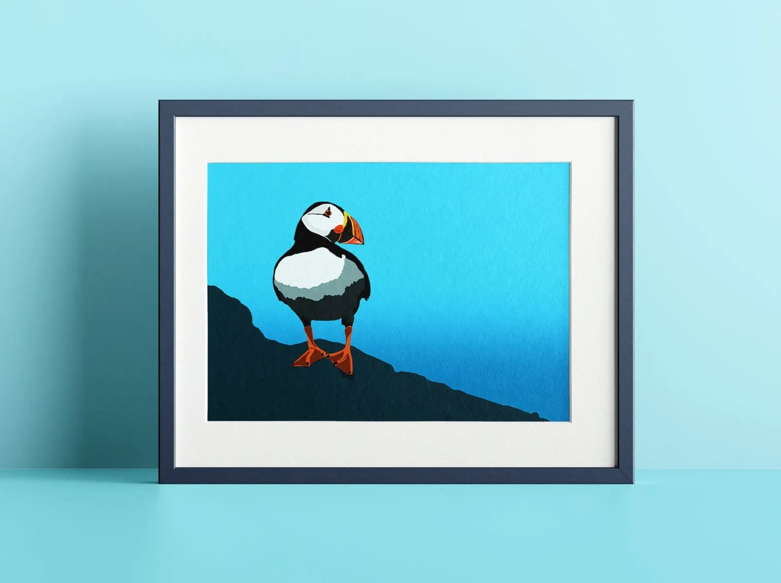 A mock up of a print in a frame against a light blue background. The print is of a puffin stood on dark green ground, against a blue gradient background.