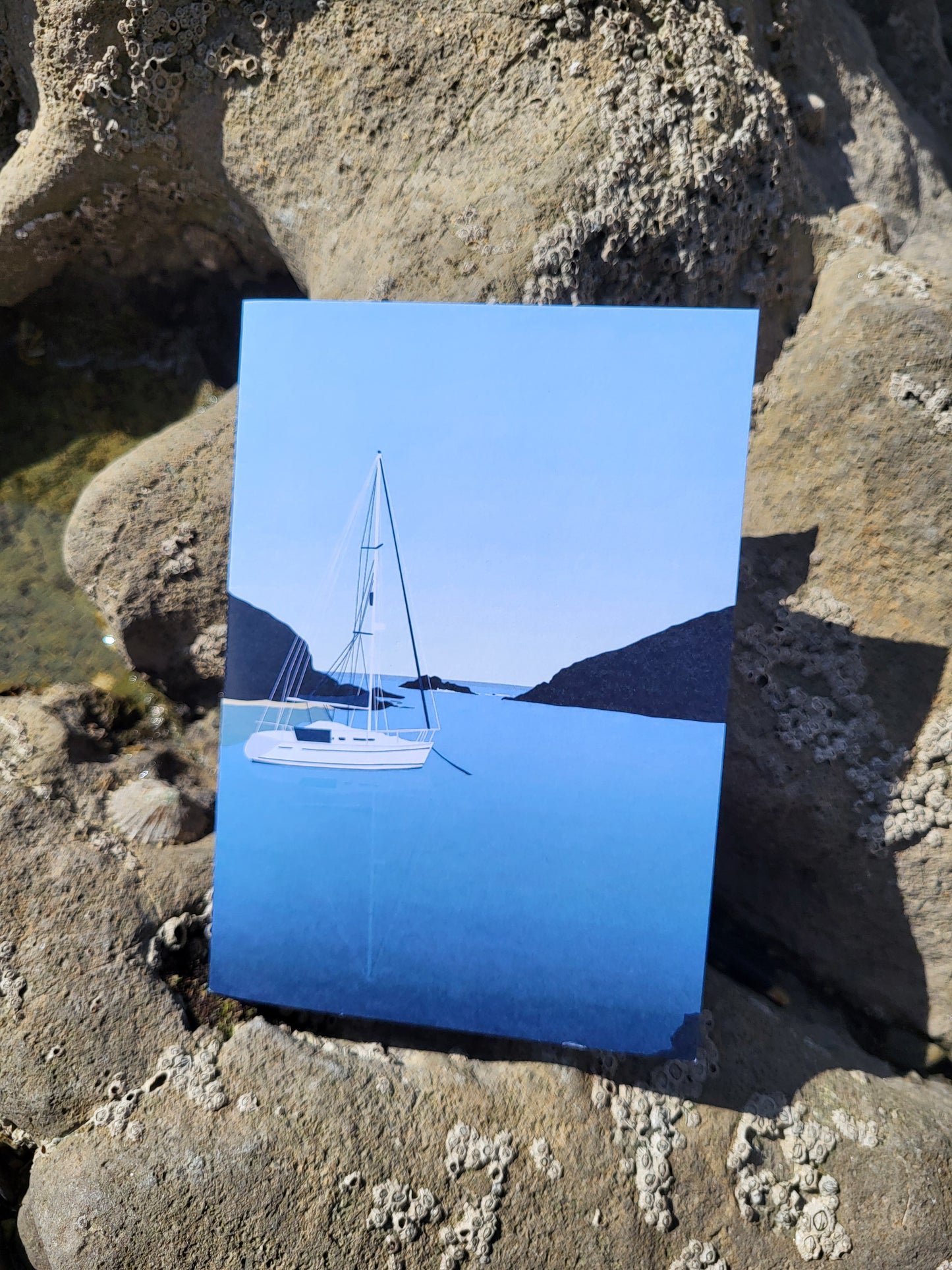 A photo of a greeting card with a boat design on the front, pictured on rocks on the beach, in front of a rockpool.