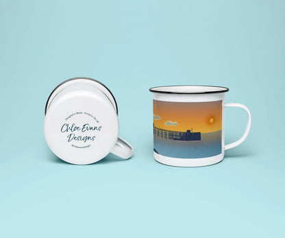 A white enamel mug from two perspectives: one of the mug on its side showing the branded base stamp on the bottom of the mug, and the other of the mug upright showing Penarth pier at sunrise; all on a light blue background.