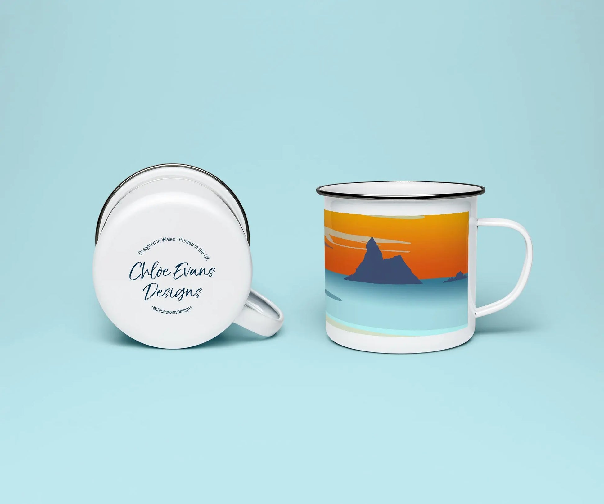 A mock up of a white enamel mug from two perspectives; one of the mug on its side looking at the bottom of the mug and the branded base stamp, and the other of the mug upright showing the print of an orange and blue beach scene, showing Church Rock and Broad Haven South at sunset, all on a light blue background.