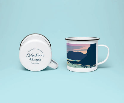 A white enamel mug from two perspectives: one of the mug on its side showing the branded base stamp on the bottom of the mug, and the other of the mug upright showing the pink, orange and blue print of Caswell beach at sunset; all on a light blue background.