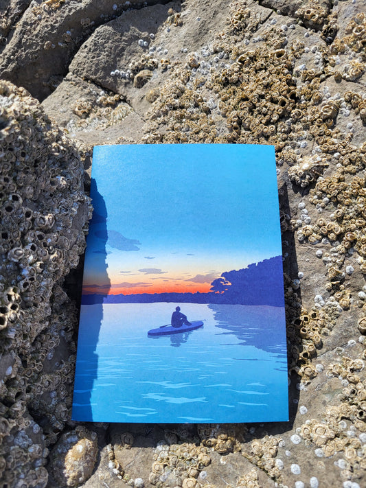 A photo of a greeting card with a paddleboarder looking at the sunset design, pictured on barnacle covered rocks at the beach.