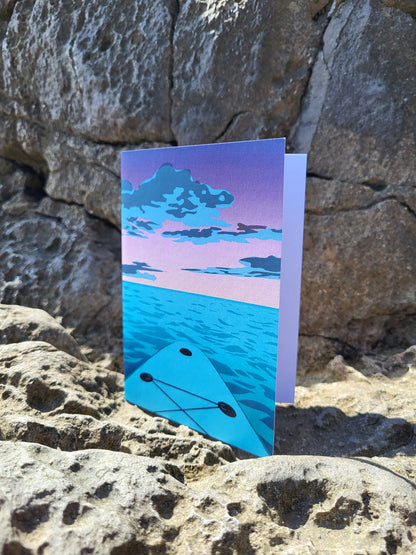 A greeting card with a purple, pink and blue paddle board sunset design, pictured on rocks at the beach.