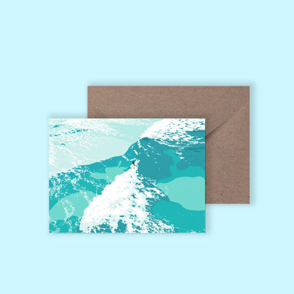 A mockup of a greeting card with an illustration of a surfer riding a wave on the front, with a brown envelope behind on a light blue background.