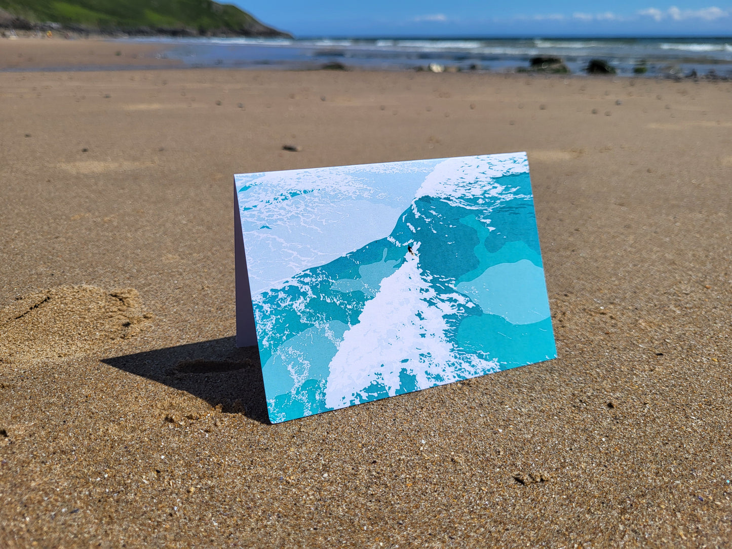 An A6 greeting card with a surf inspired design, pictured on the beach.