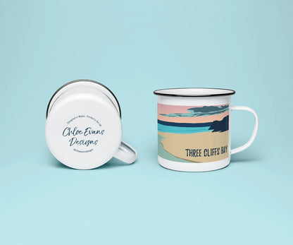 A white enamel mug from two perspectives: one of the mug on its side showing the branded base stamp on the bottom of the mug, and the other of the mug upright showing the blue, pink and orange sunset illustration of Three Cliffs bay at sunset; all on a light blue background.