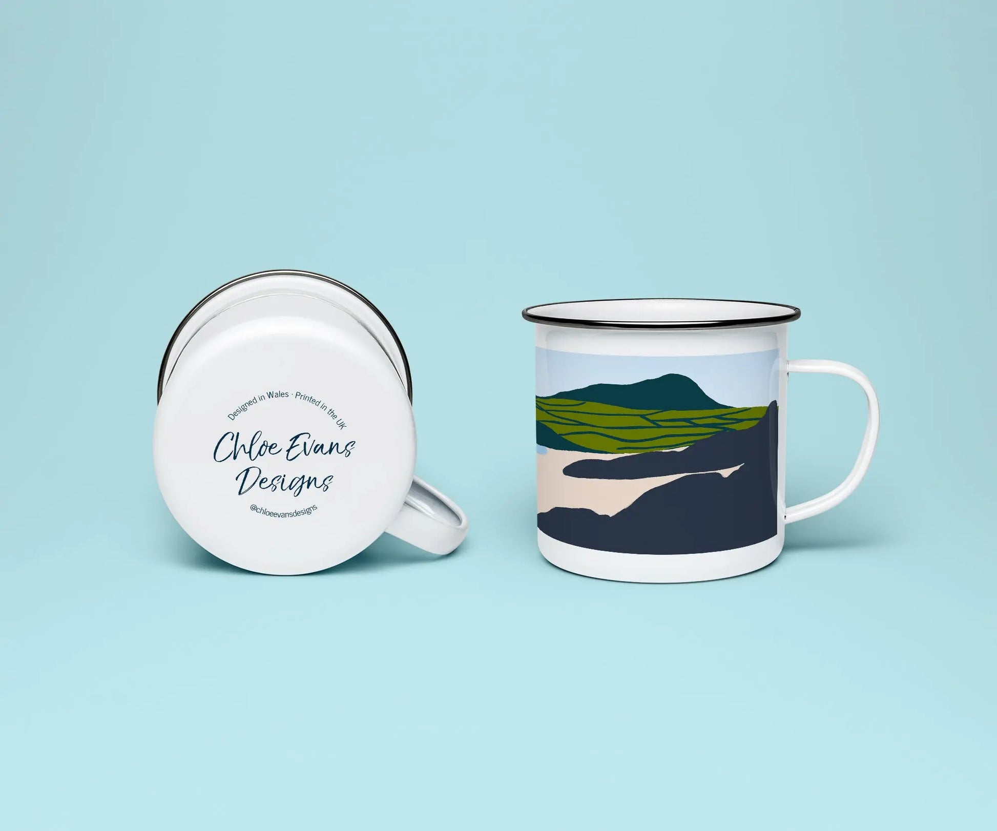 A mock up of a white enamel mug from two perspectives; one of the mug on its side looking at the bottom of the mug and the branded base stamp, and the other of the mug upright showing the print of an green, grey, and blue beach scene, showing Whitesands beach, all on a light blue background.